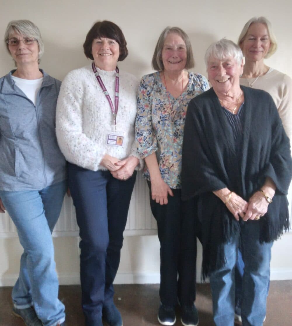 Five older women standing together indoors, smiling at the camera against a light-coloured wall. - Home Instead