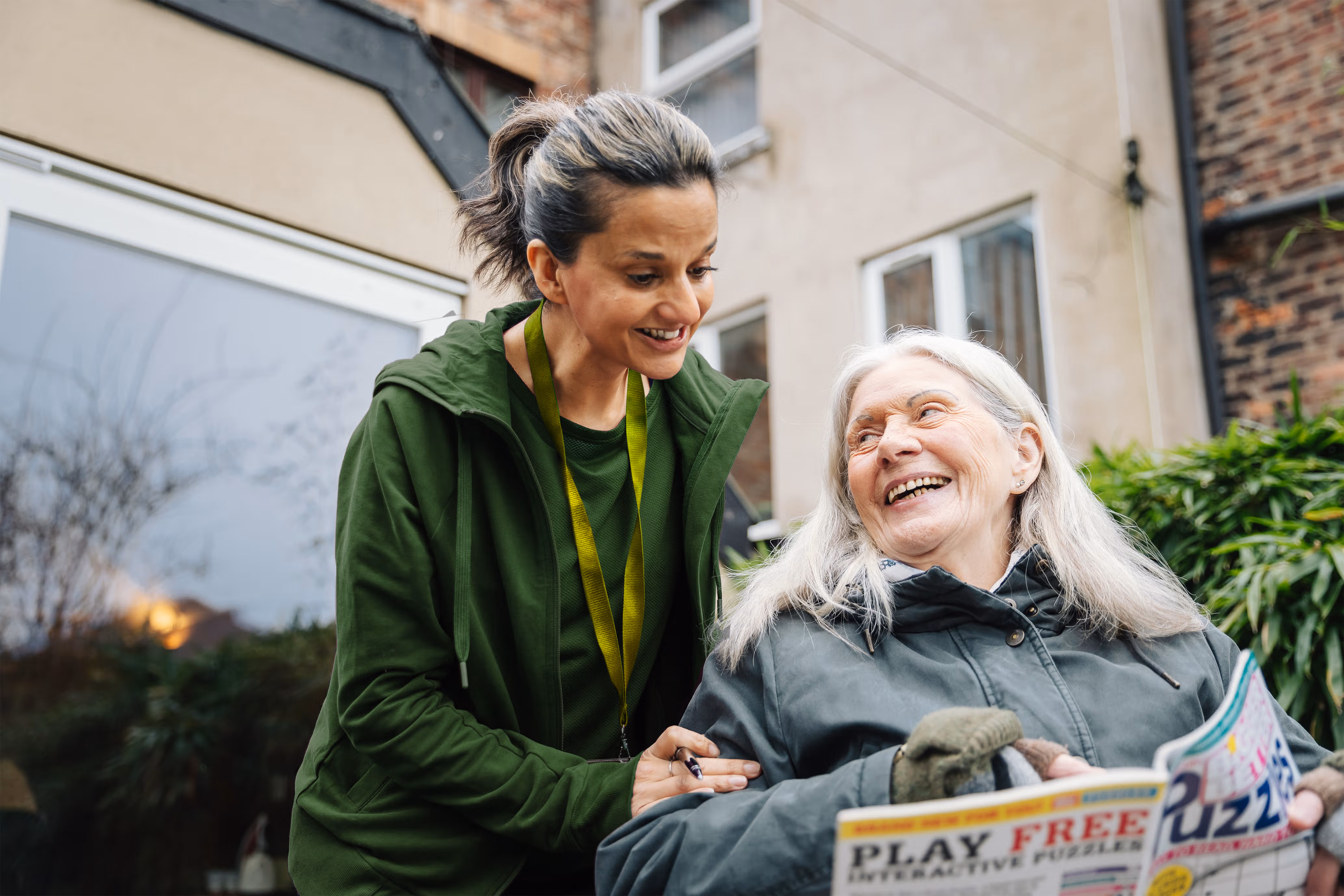 A younger woman smiles at an older woman holding a puzzle book while sitting outside in a garden. - Home Instead