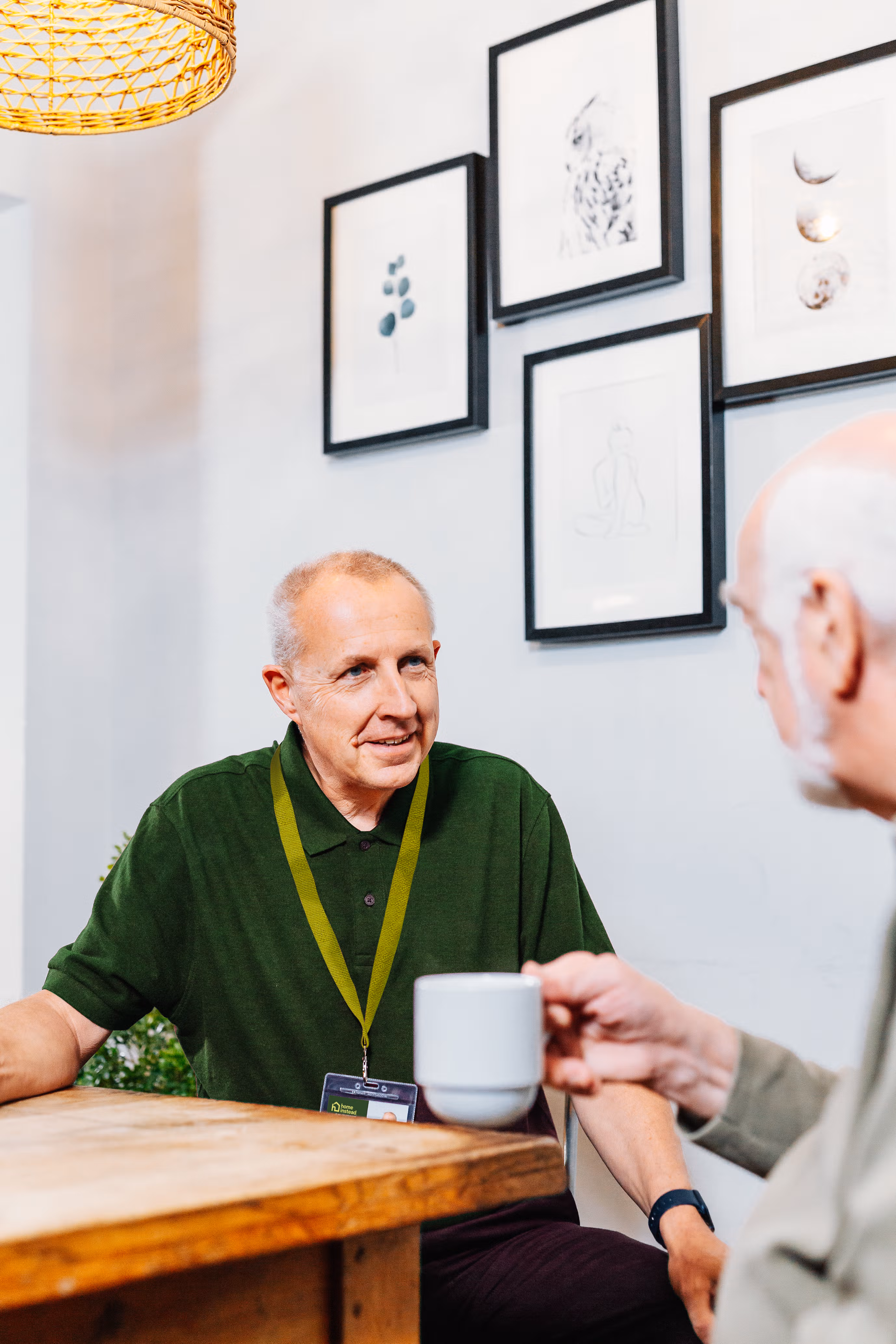 Two older men talking at a table, one holding a mug, with framed art on the wall behind them. - Home Instead