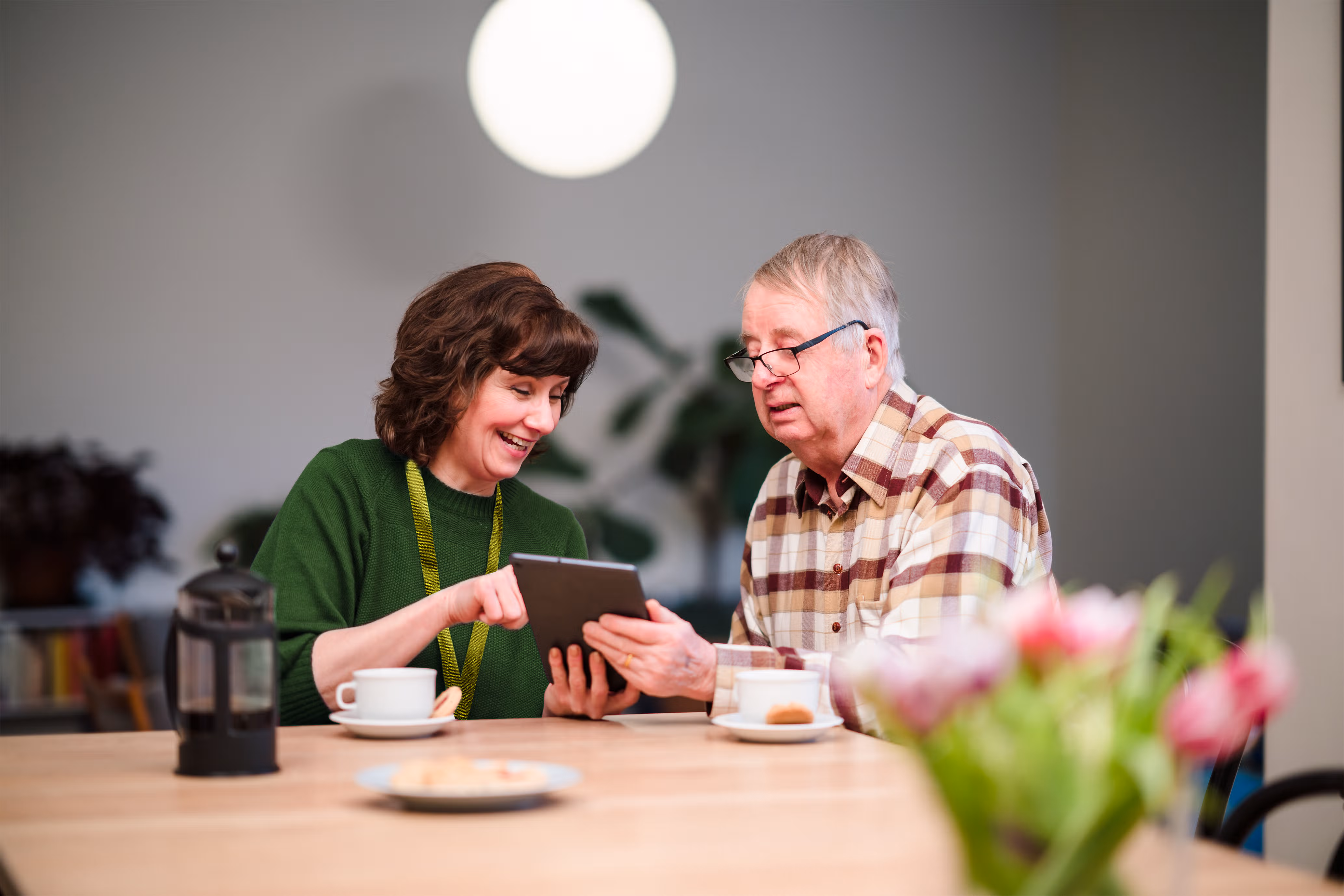 Smiling woman and elderly man sit at a table, looking at a tablet together in a cosy room. - Home Instead