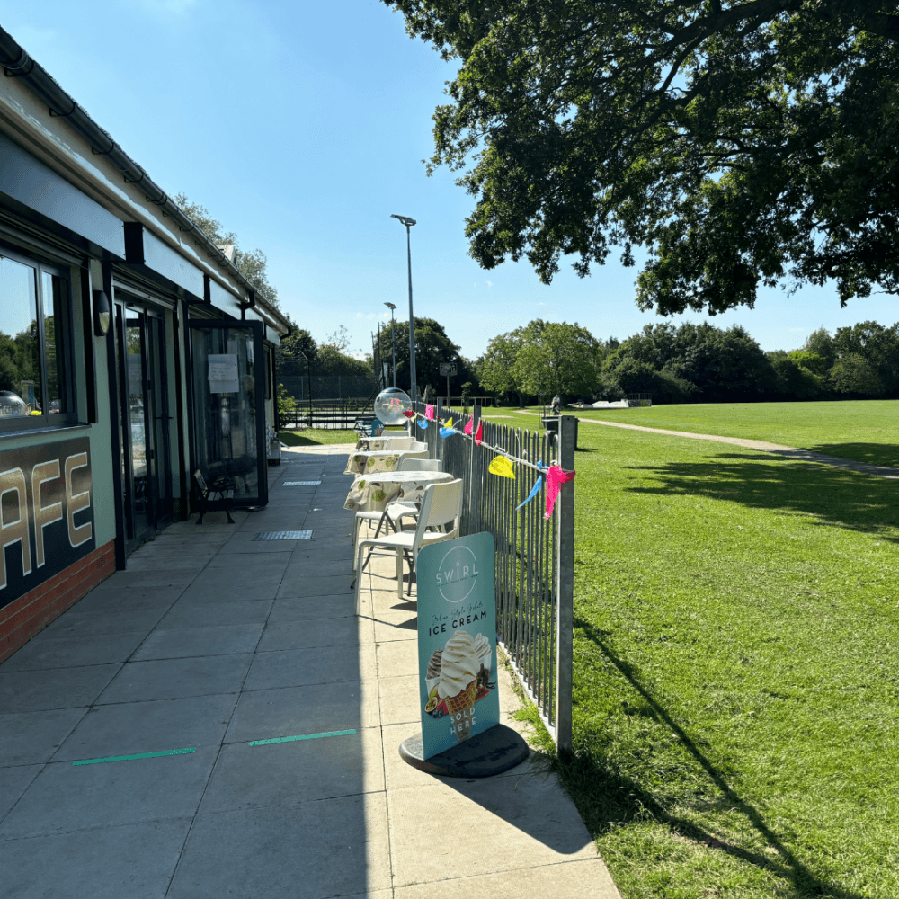 Outdoor café with a sidewalk sign advertising ice cream, tables with umbrellas, and a view of a sunny park. - Home Instead