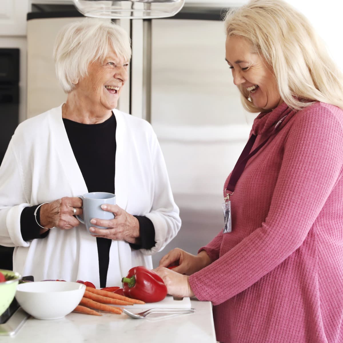 Two elderly women in a kitchen, one holding a mug, the other slicing a red bell pepper. Both are smiling and talking. - Home Instead