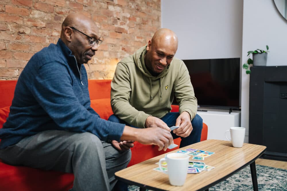 Two men are sitting on a sofa playing a card game. A coffee table with two mugs is in front of them. - Home Instead
