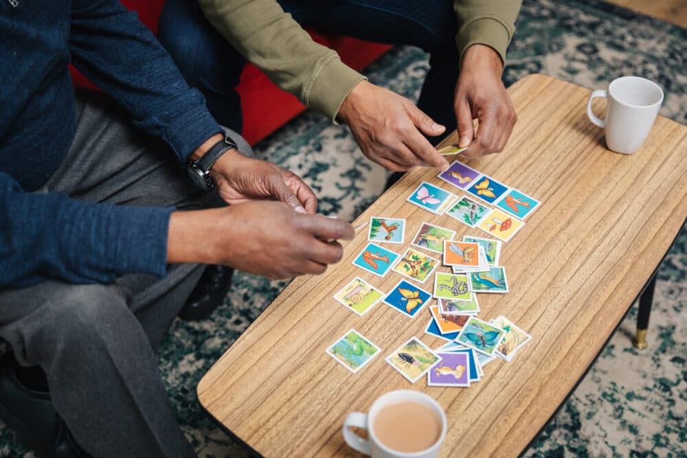 Two people playing a card game on a wooden table, with two white mugs nearby. - Home Instead