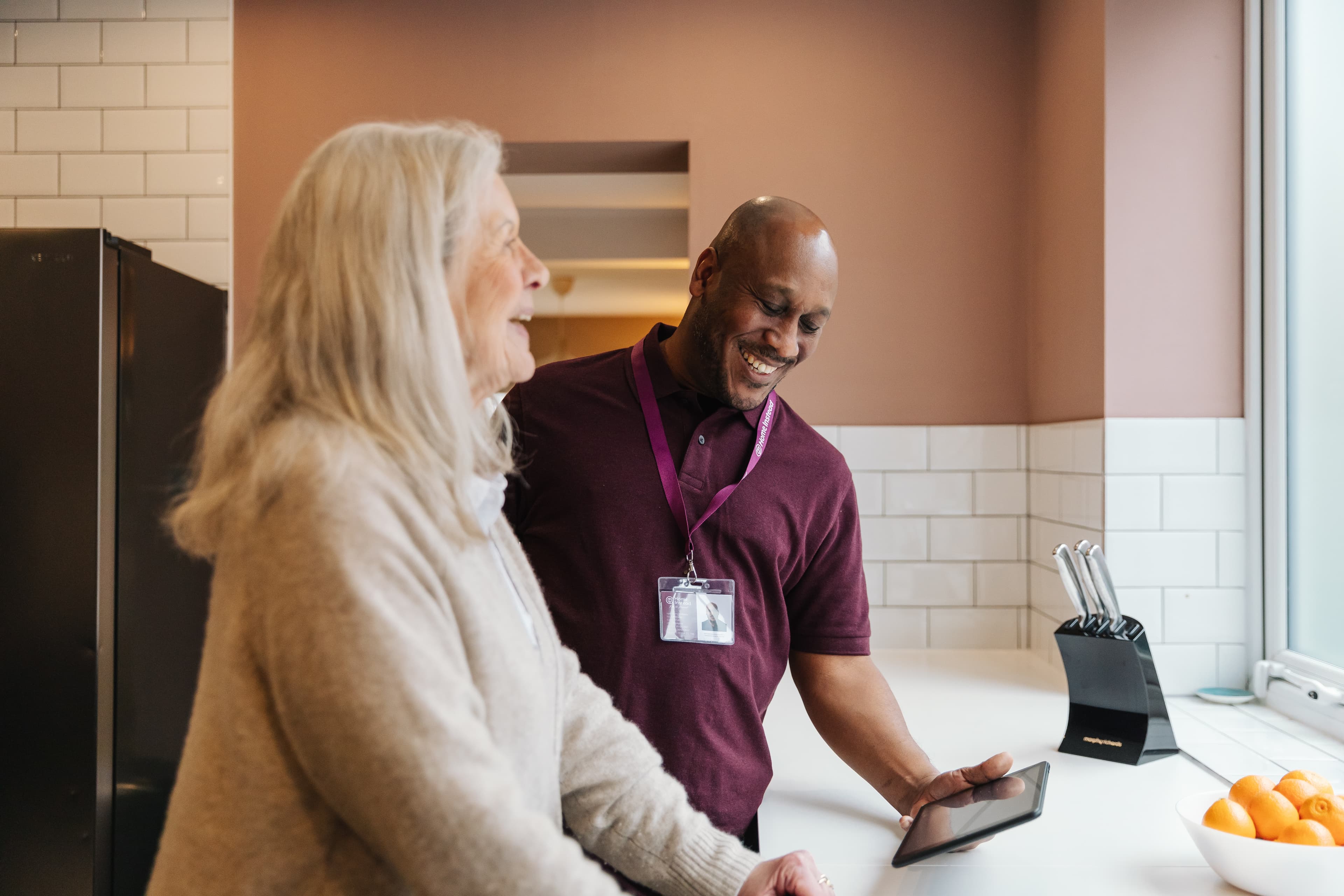 An elderly woman and a man with a tablet smile in a kitchen setting. - Home Instead
