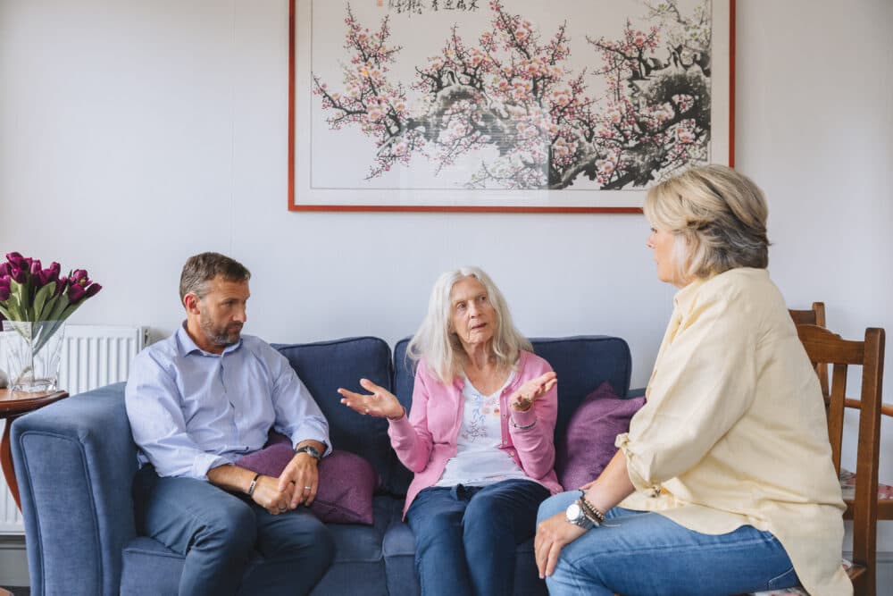 Three people seated on a couch and a chair, having a conversation, with a painting of flowers on the wall behind them. - Home Instead