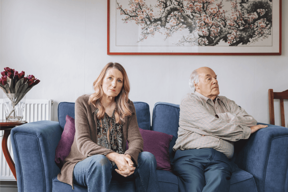 A woman and an older man sit apart on a blue couch, appearing distant. A painting and flower vase are in the background. - Home Instead