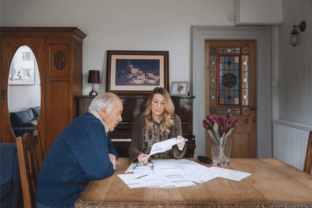 An elderly man and a woman review documents together at a table in a cozy room with stained glass and flowers. - Home Instead