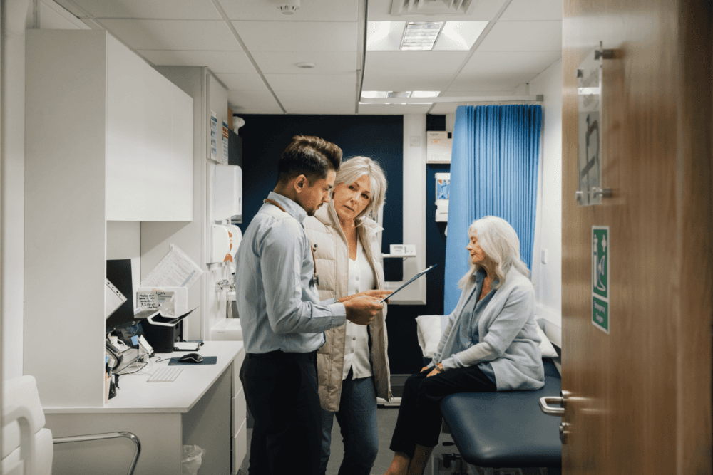 A doctor discusses a patient's chart with a woman as an elderly woman sits on an examination table in a medical room. - Home Instead