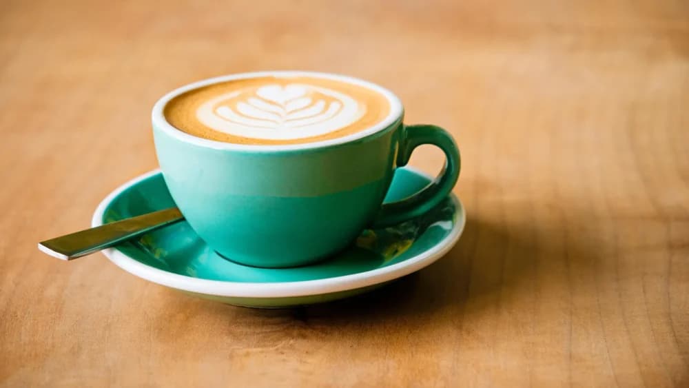 A teal cup of coffee with latte art and a spoon on a matching saucer, placed on a wooden table. - Home Instead
