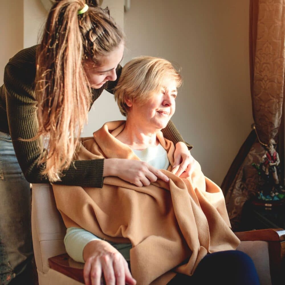 A young woman gently hugs an older woman sitting in a chair, both smiling warmly. - Home Instead