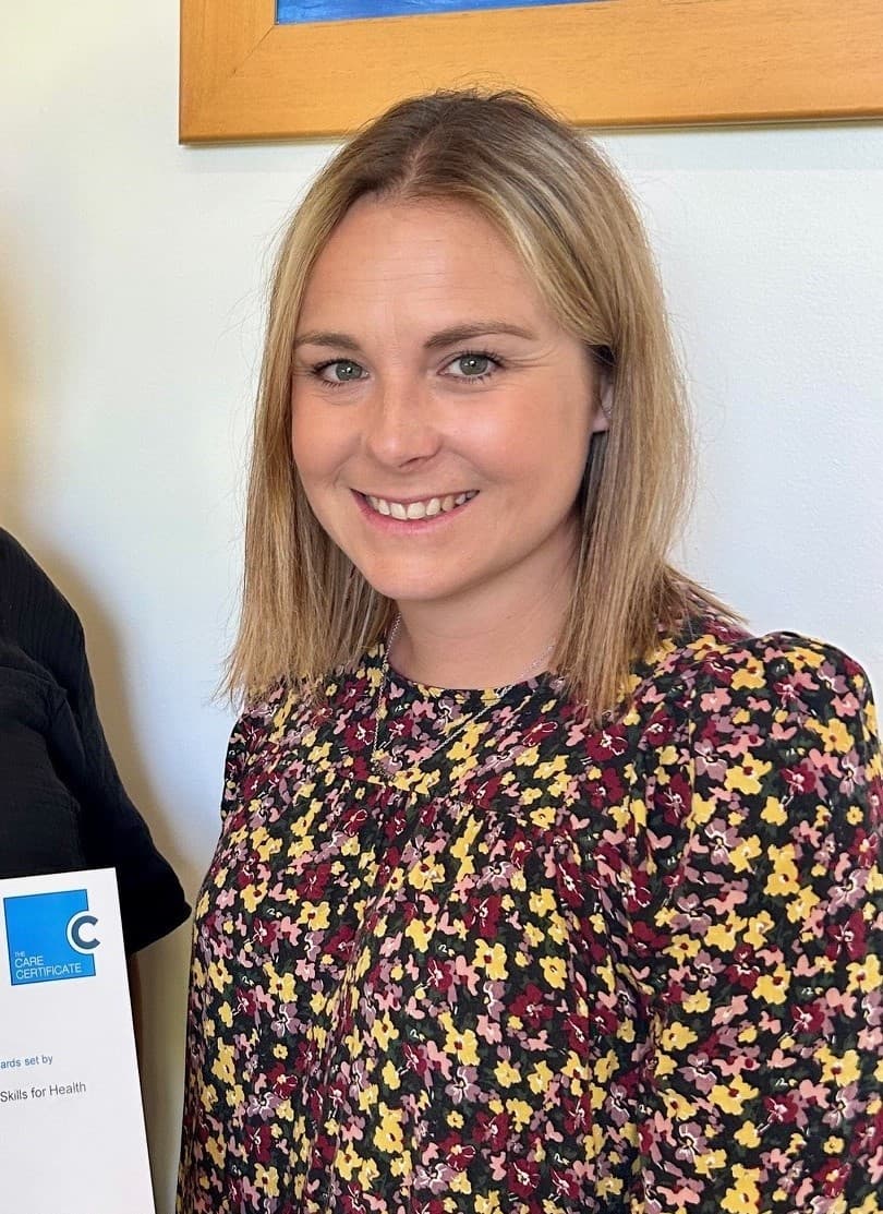Smiling woman with blonde hair wearing a floral blouse, standing indoors near a partially visible certificate. - Home Instead