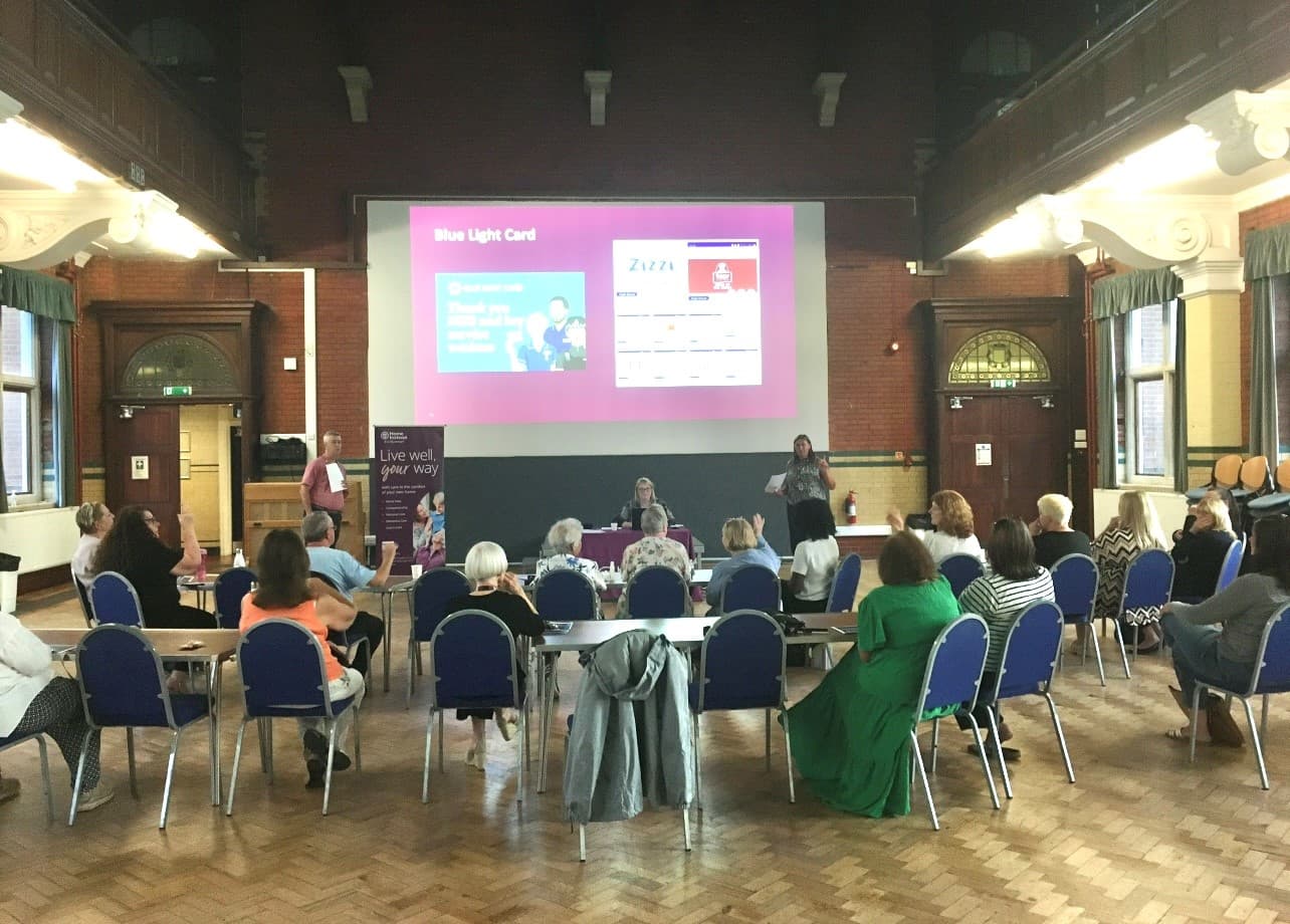 Group meeting in a hall with a presentation screen. People seated in a semicircle around tables. - Home Instead
