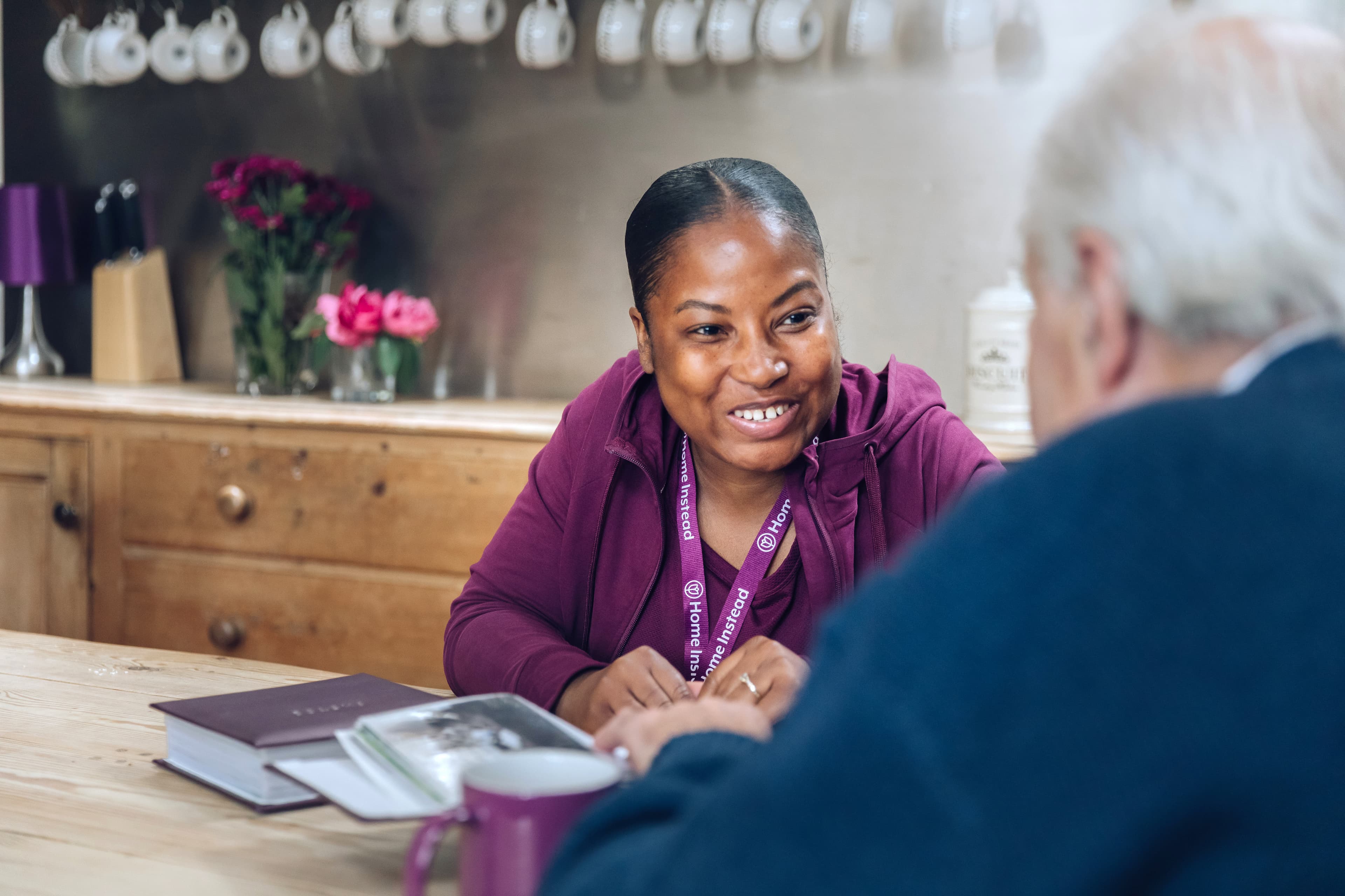 A woman in a purple uniform smiling and talking to an older man at a wooden table with flowers in the background. - Home Instead