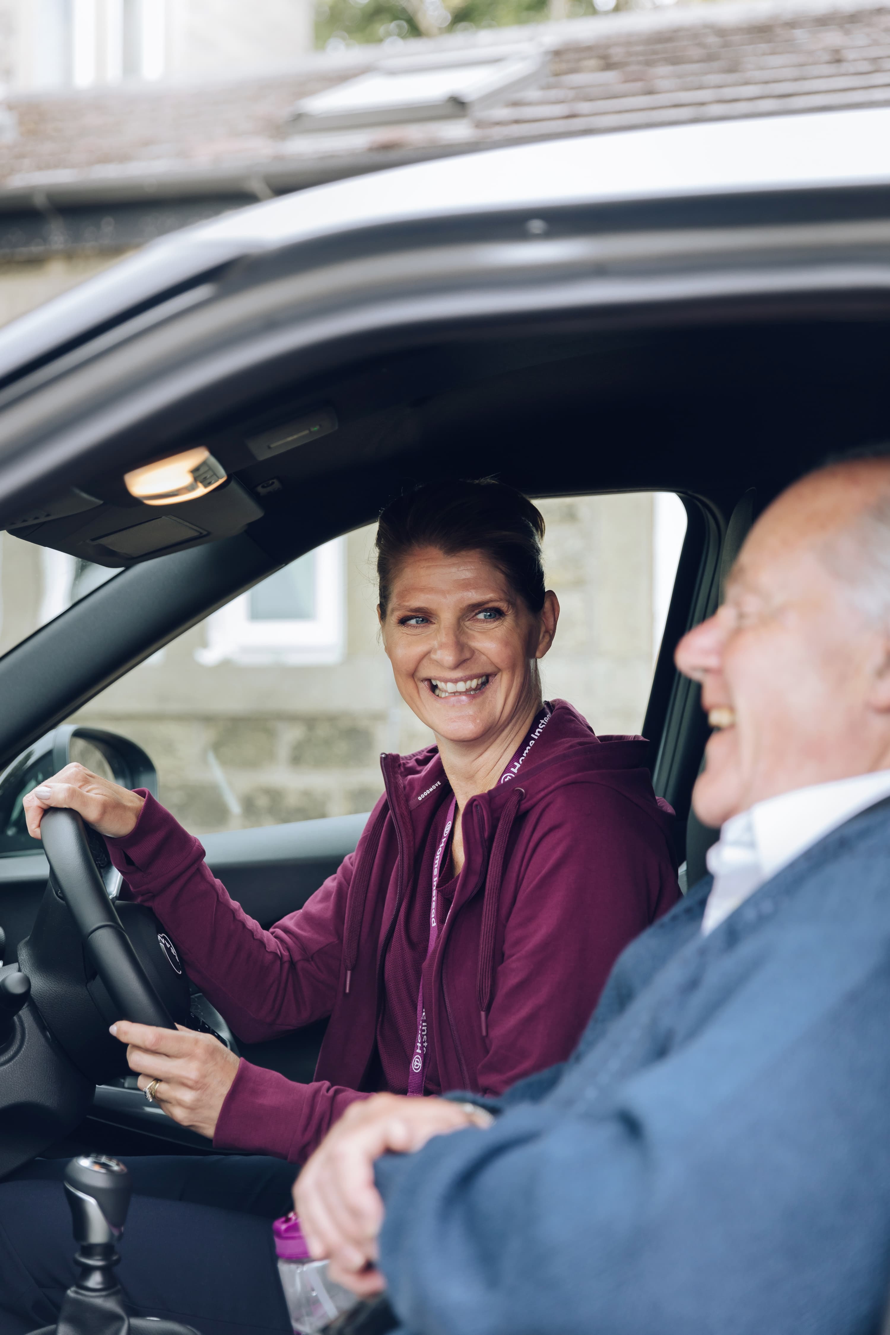 A woman in a maroon jacket smiles while sitting in a car, next to an elderly man who is laughing. - Home Instead