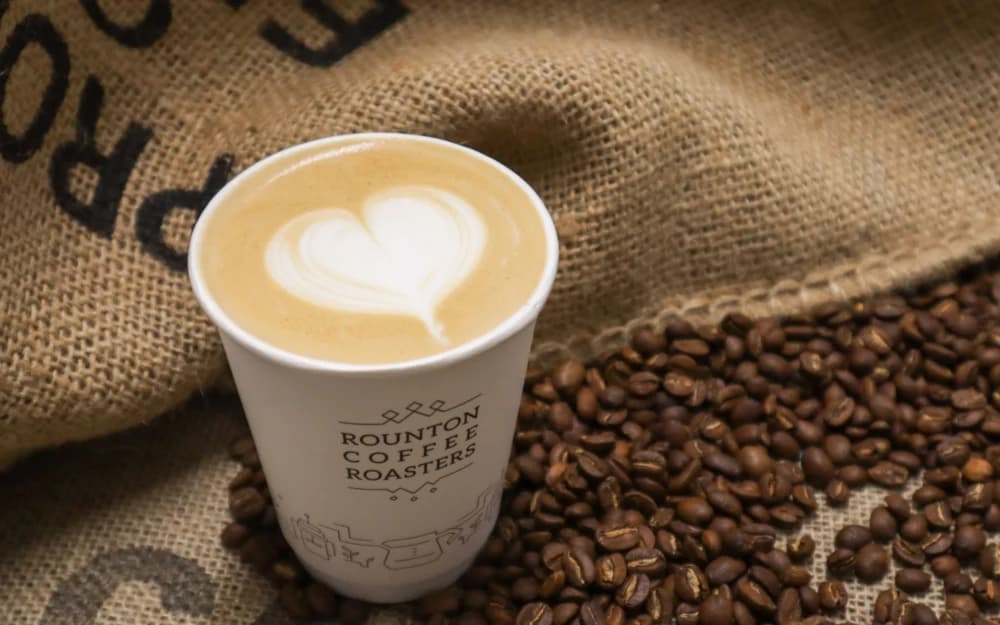A cup of latte with a heart design, surrounded by coffee beans and a burlap sack background. - Home Instead