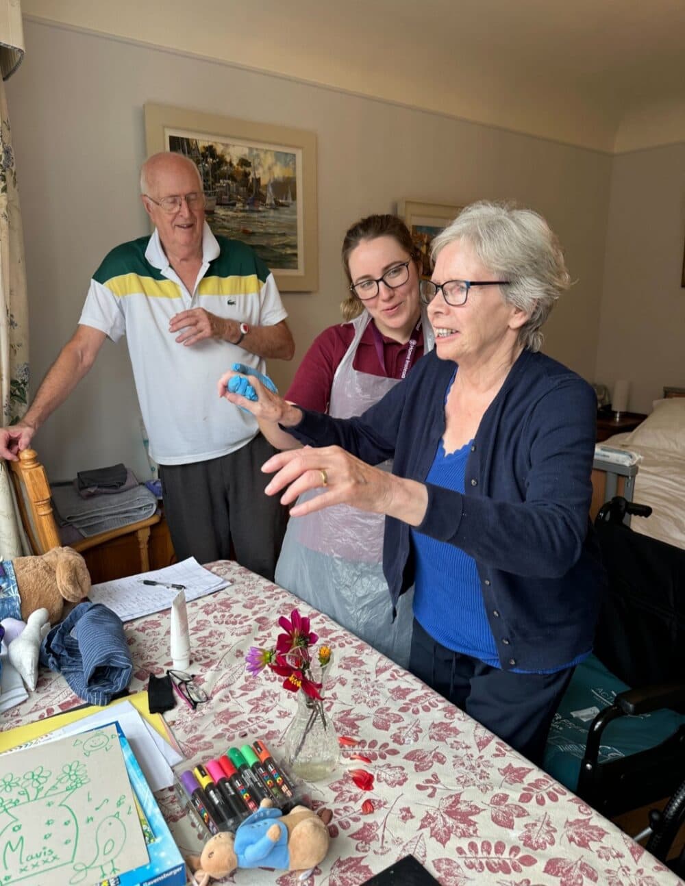 Three people smiling and playing with crafts at a table, one seated, two standing, in a cozy room. - Home Instead