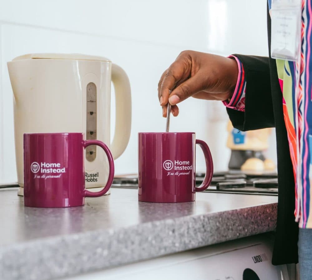 Person stirring a purple mug next to a kettle on a kitchen counter. - Home Instead