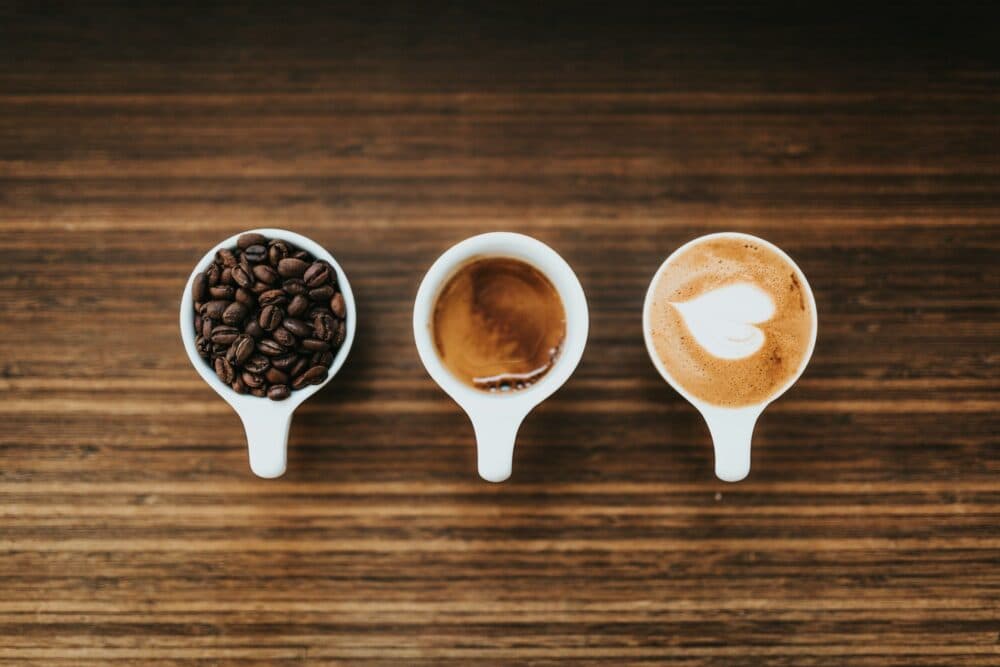 Three white cups on a wooden table with coffee beans, espresso, and a cappuccino with latte art. - Home Instead