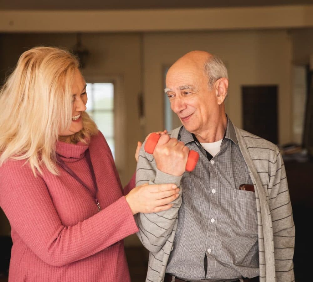 New Ageing Index Report, image shows woman with blonde hair, wearing a pink long sleeved top and a lanyard, assisting older man wearing shirt and cardigan to lift a small dumbbell in a home setting.