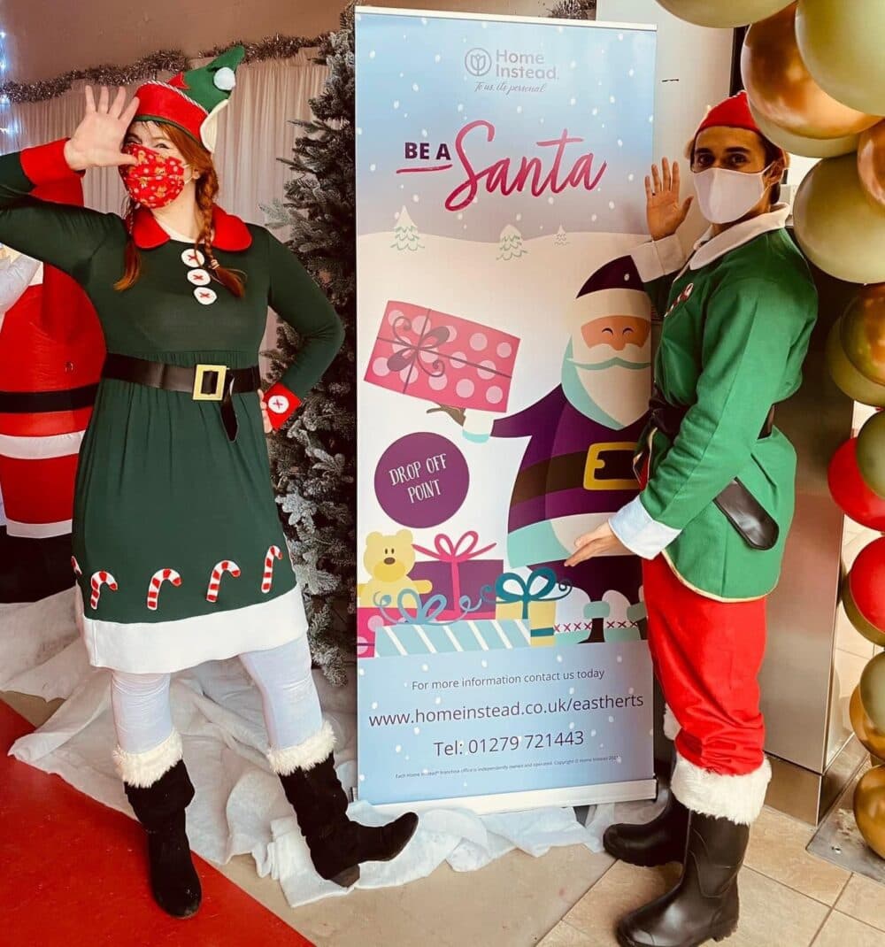 Two people dressed as elves posing next to a "Be a Santa" banner with Christmas gifts and decorations around. - Home Instead