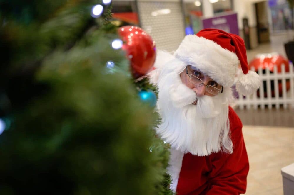 Santa Claus playfully peers from behind a Christmas tree, adorned with lights and ornaments, in a festive setting. - Home Instead