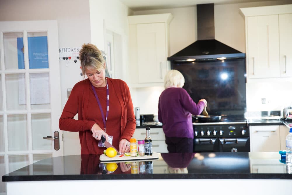 Two women prepare food in a modern kitchen; one chops a pepper, the other cooks at the stove. - Home Instead