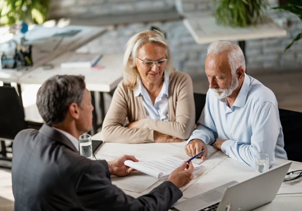 An older couple reviews documents with a suited man at a table in an office setting. - Home Instead