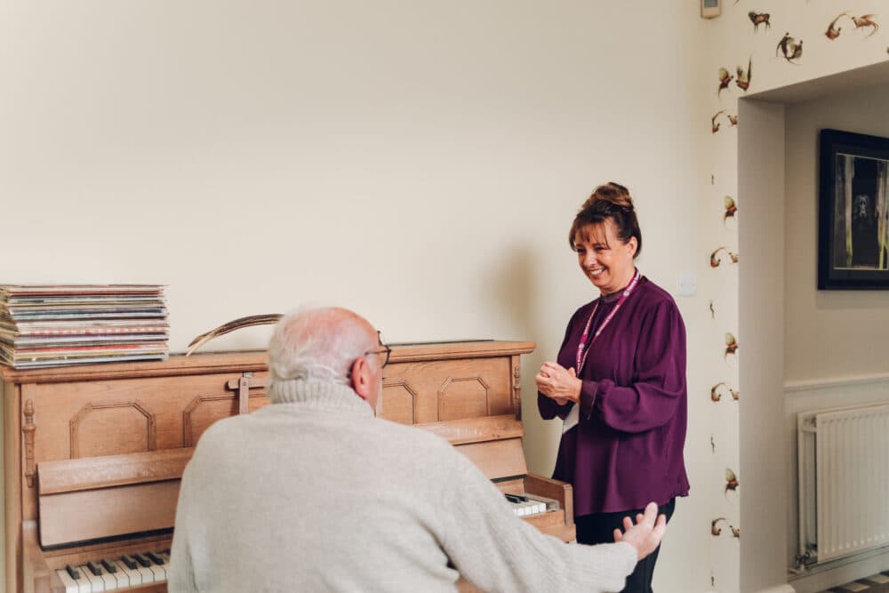 An elderly man plays piano while a woman in a purple blouse stands nearby, smiling warmly. - Home Instead