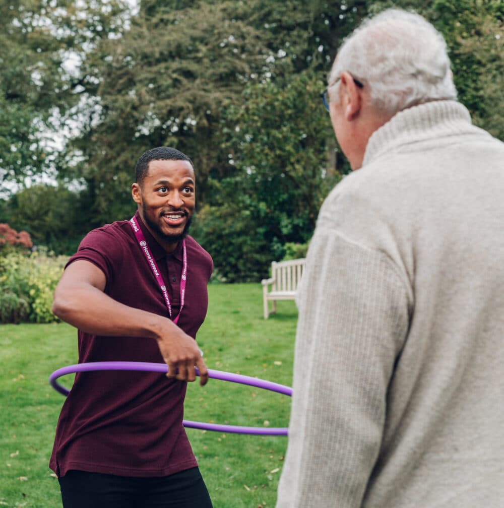 A man in a burgundy shirt hula-hoops while smiling at a person in glasses and a sweater in a park. - Home Instead