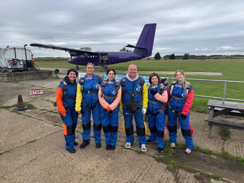 Six people in blue jumpsuits stand smiling in front of a purple airplane on a cloudy day. - Home Instead