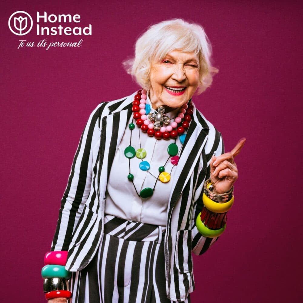 Elderly woman in colorful jewelry and striped outfit smiling, with "Home Instead" logo. - Home Instead