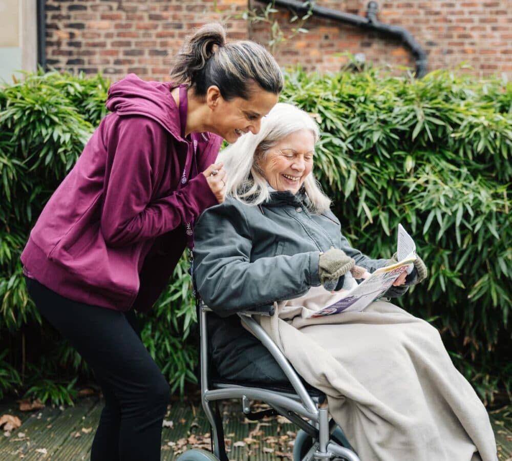 A senior woman with white hair sitting down while looking at a puzzle with a young carer smiling and standing and wearing a sweater outside their garden