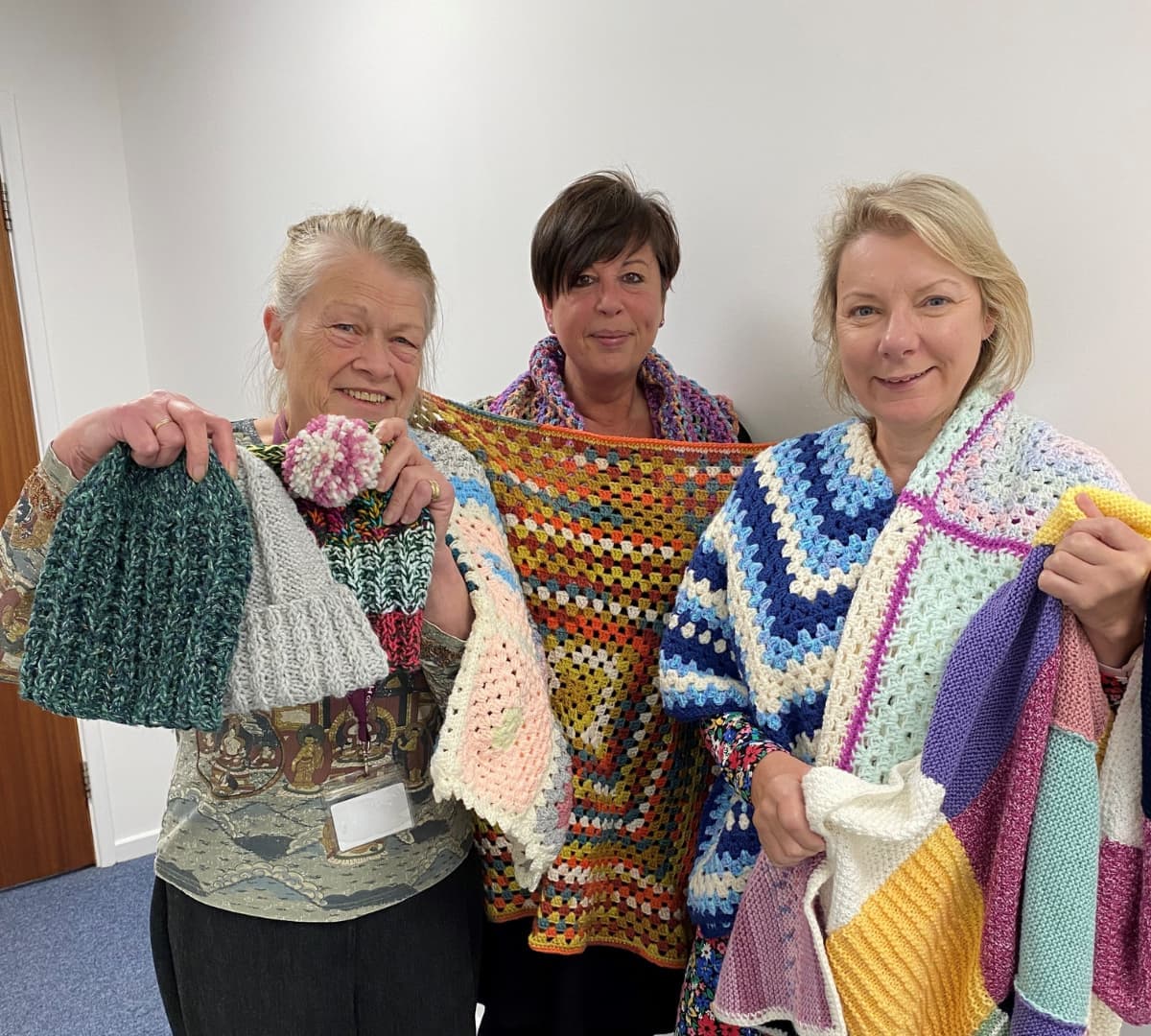 Three women holding colorful knitted items and blankets, smiling in a room. - Home Instead