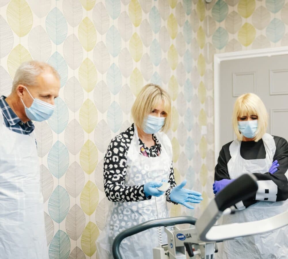 Two women and one man wearing masks, gloves and aprons looking at a chair lift hoist inside a room