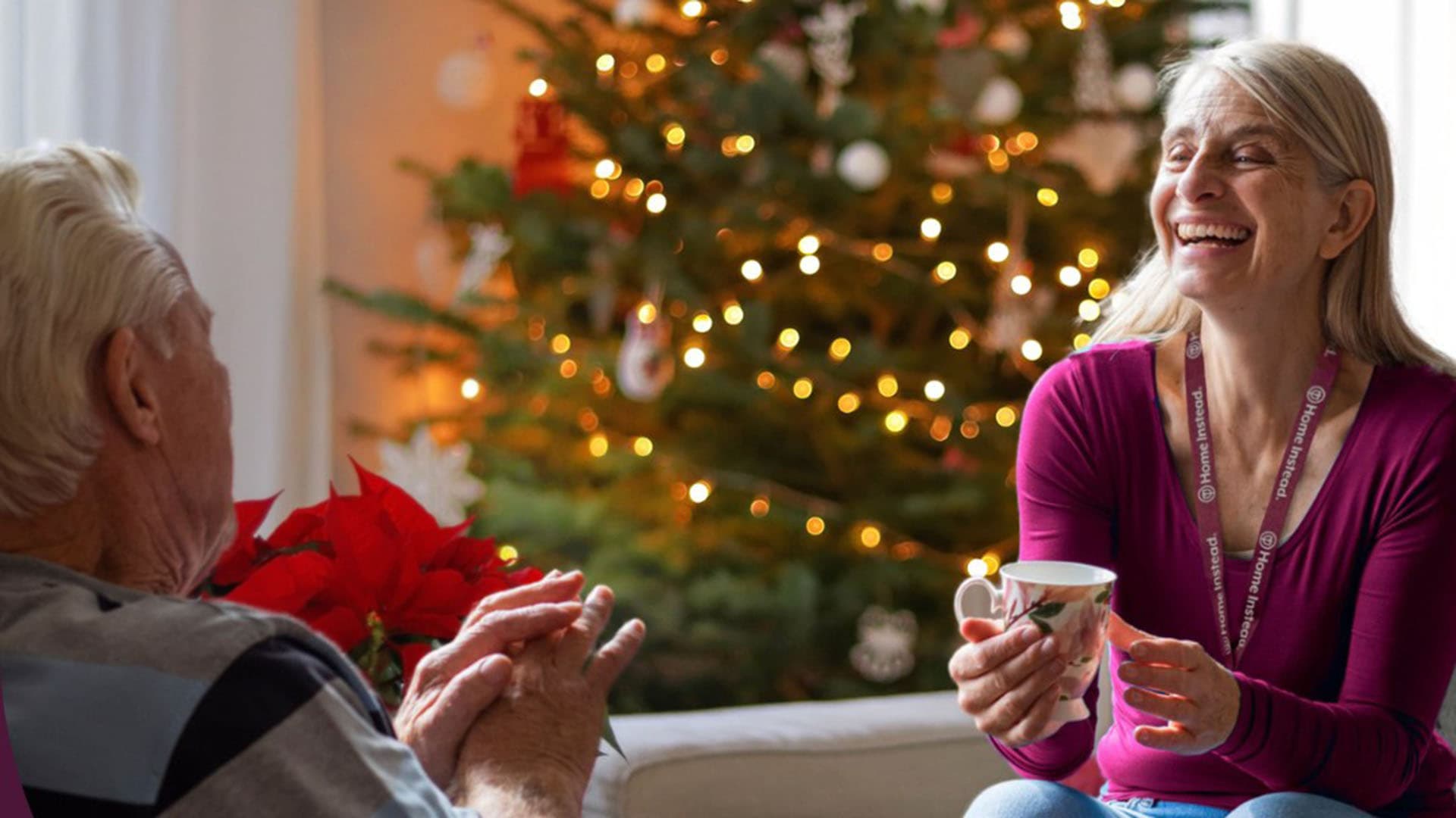 An older man and woman smiling and talking by a lit Christmas tree, holding a cup and sitting indoors. - Home Instead