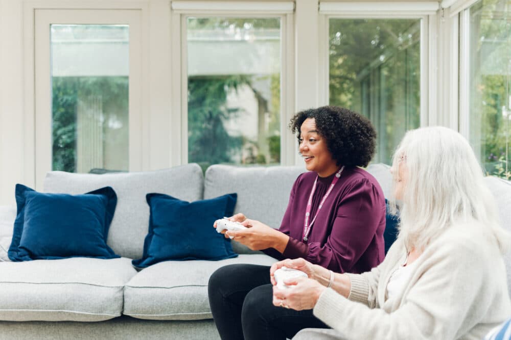 Two women sitting on a couch playing video games together in a bright living room with blue cushions. - Home Instead