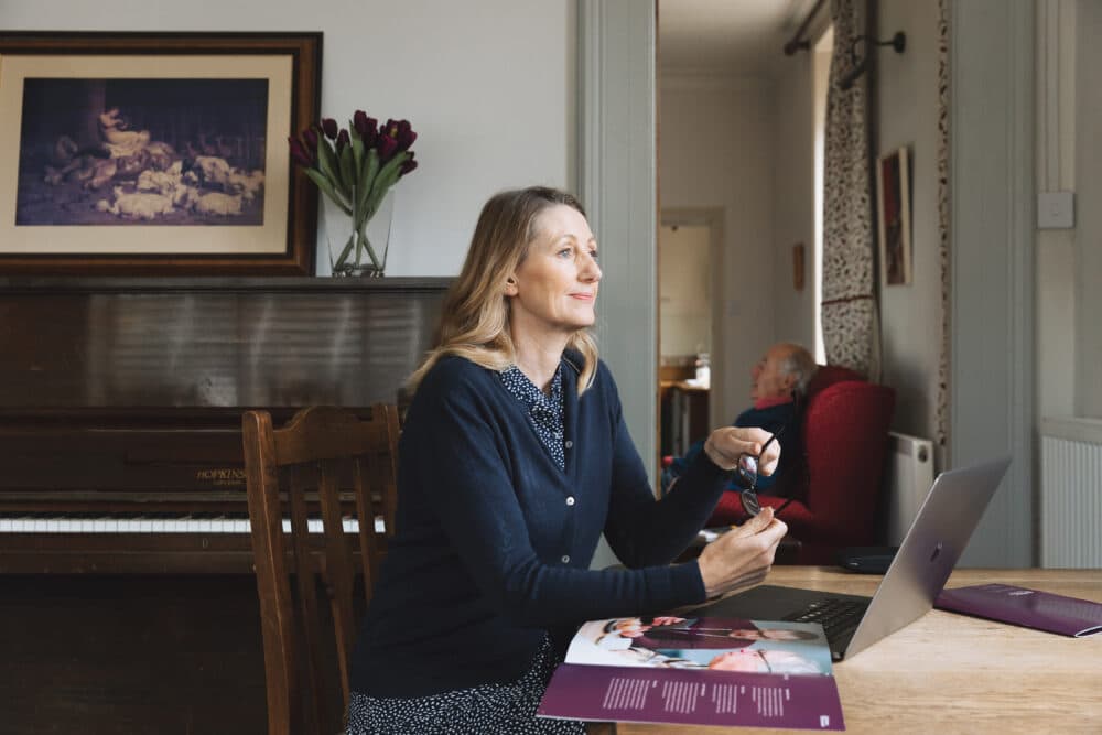 Woman sitting at a table with a laptop, holding glasses, with an older man in the background. - Home Instead
