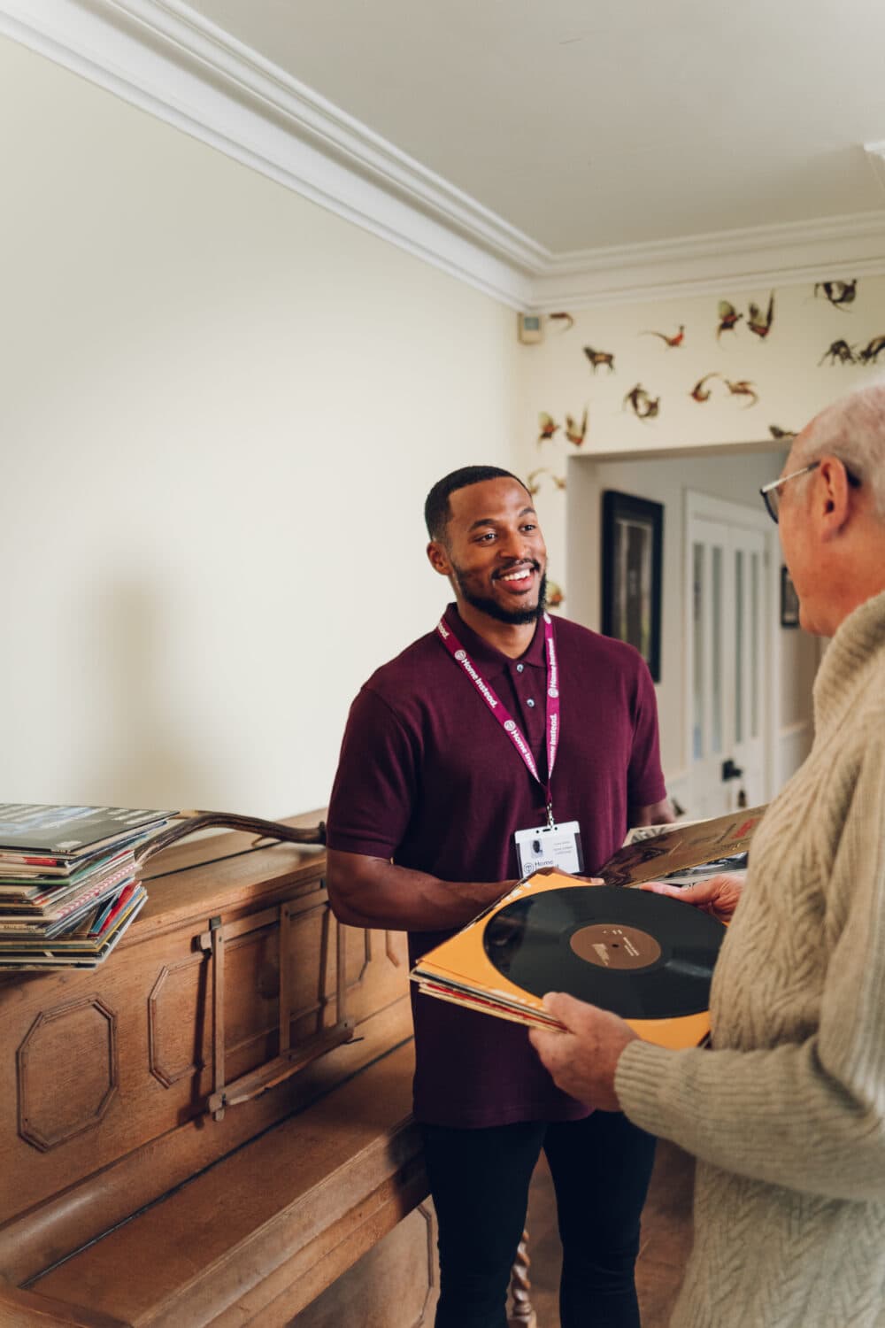 A man with a lanyard smiles while handing vinyl records to an older man by a piano. - Home Instead