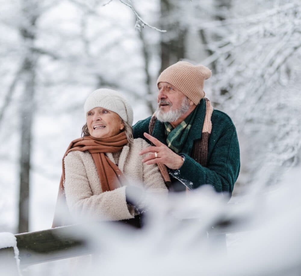 Older couple in winter clothes enjoying a snowy day outdoors, surrounded by snow-covered trees. - Home Instead