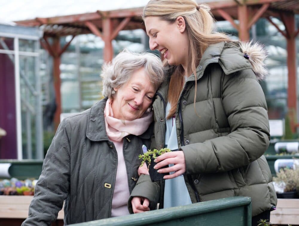 Older woman and younger woman smiling together at a garden center, holding a small potted plant. - Home Instead