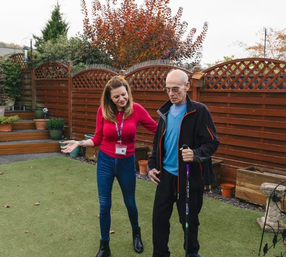 A woman assists an elderly man with a walking stick in a garden with wooden fencing and potted plants. - Home Instead