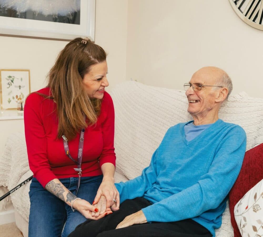 A woman smiles and holds hands with an older man sitting on a couch, both appearing happy and relaxed. - Home Instead