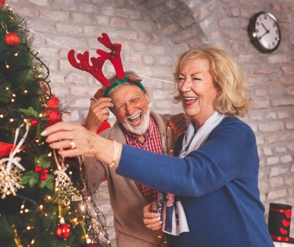 Smiling older couple decorates a Christmas tree, the man wearing reindeer antlers, in a cozy room. - Home Instead