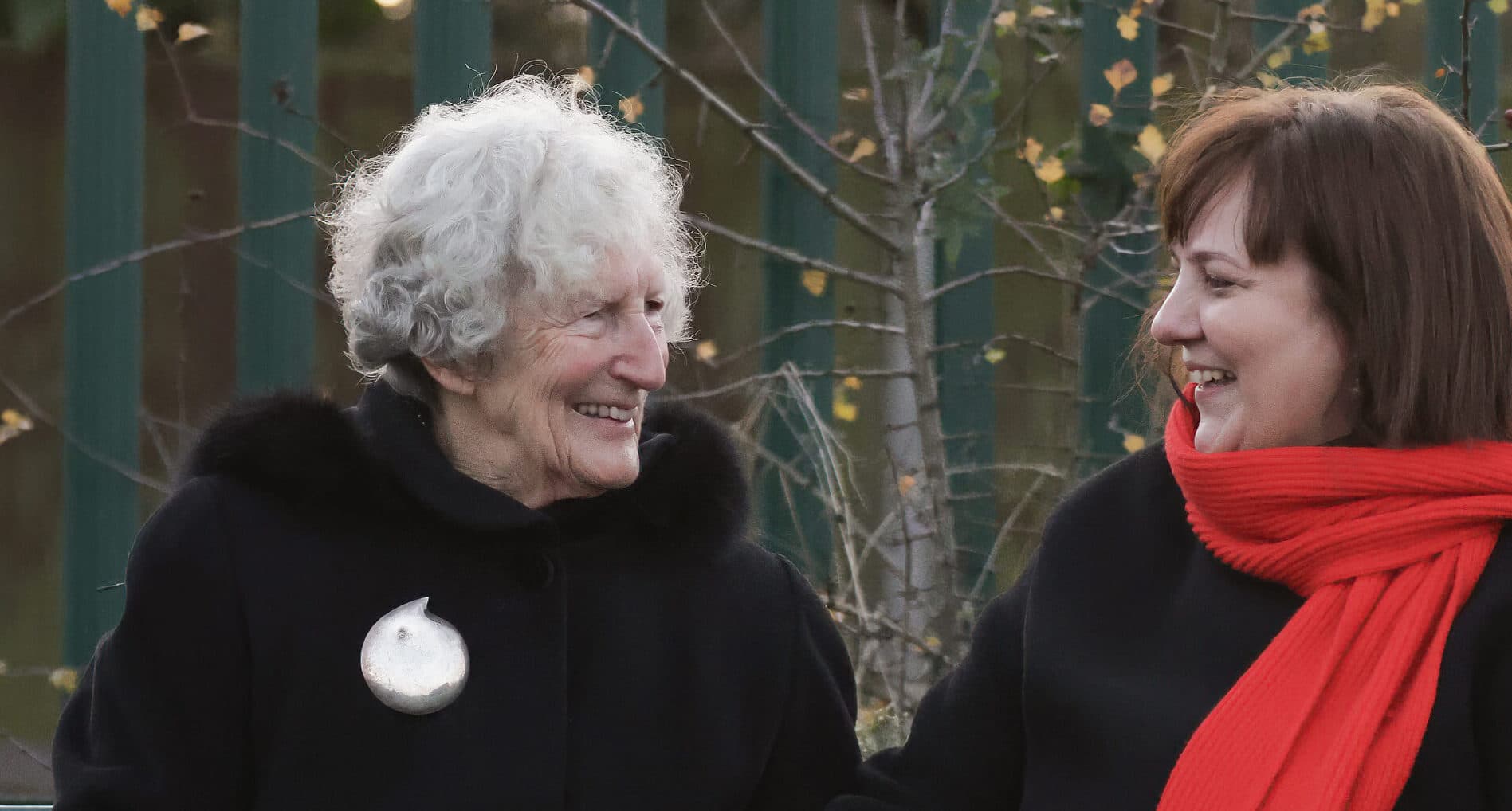 Two women sitting and smiling warmly at each other, one wearing a red scarf and the other a black coat. - Home Instead
