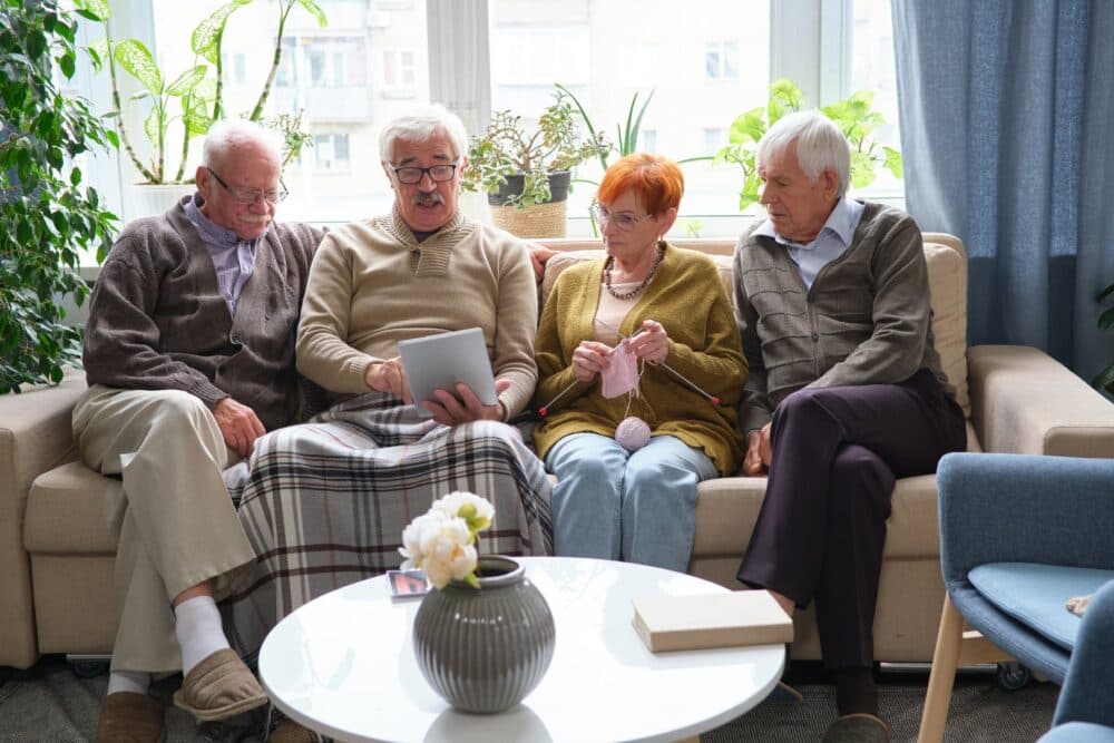 Four elderly people sit on a couch; one uses a tablet, another knits, with plants and books nearby. - Home Instead