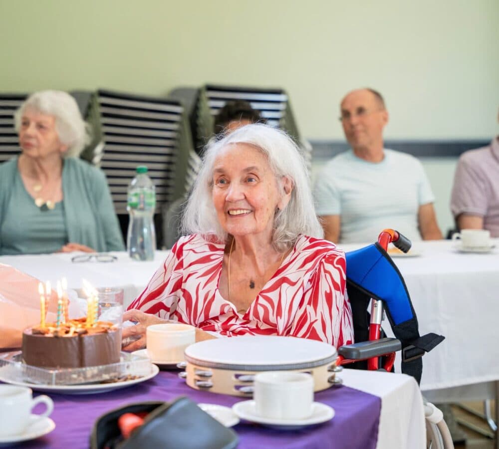 Senior woman with grey hair smiling and happy while sitting on a wheelchair with cake and candles on the table and people at her back