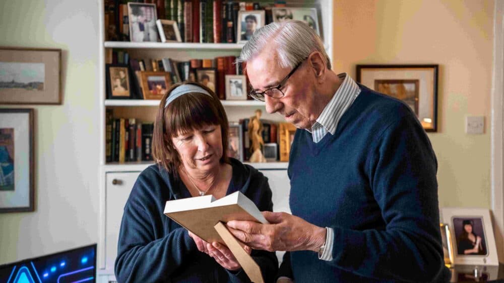 Older man and woman looking at a framed photo together in a cozy room with bookshelves and pictures. - Home Instead