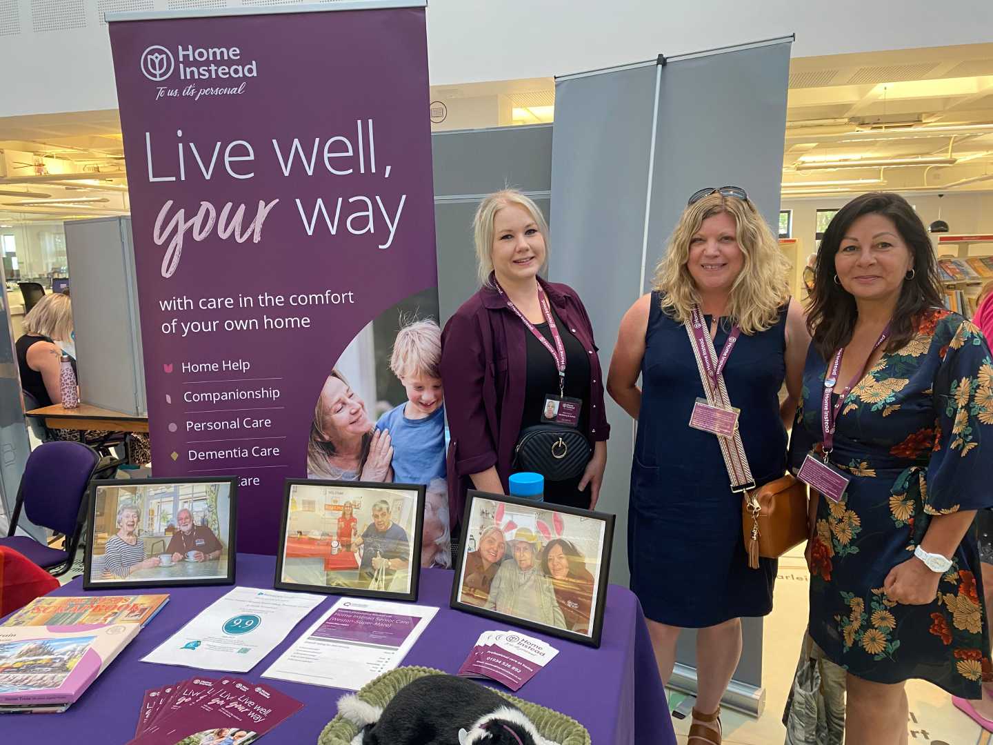 Three women stand by a Home Instead care service booth with brochures, photos, and a display banner. - Home Instead