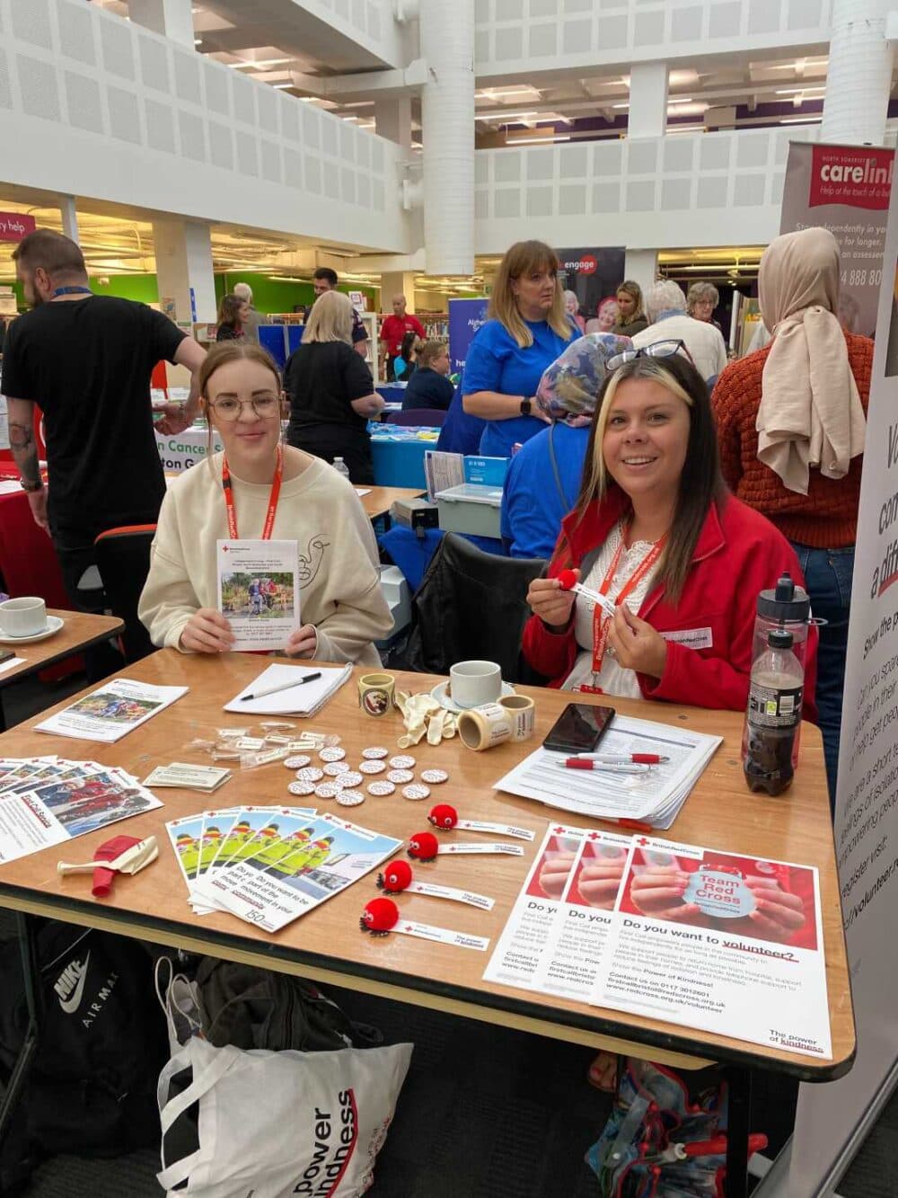Two women sit at a stall with leaflets, buttons, and flyers at a busy indoor event or fair. - Home Instead
